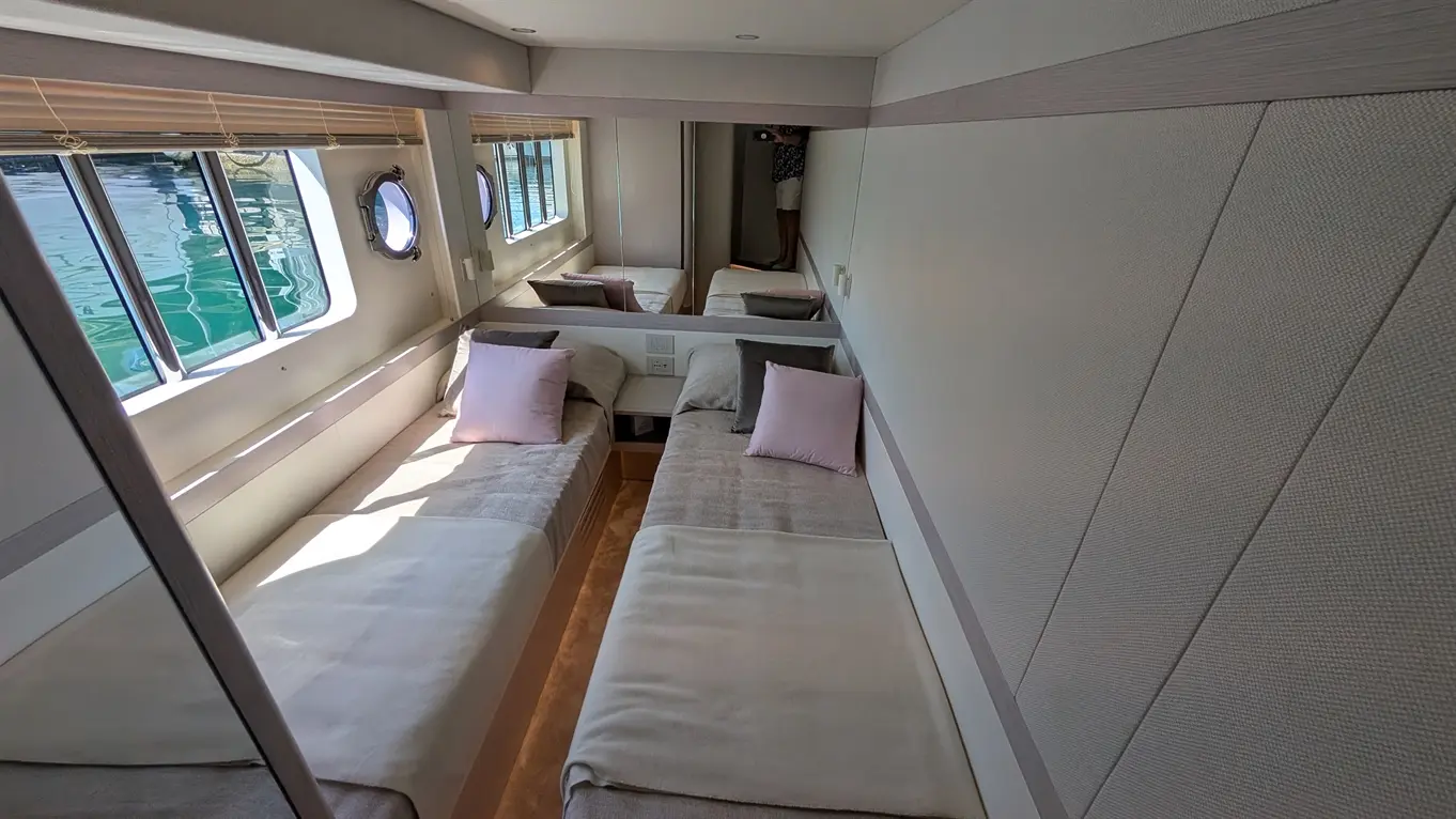 Brightly lit cabin interior with two twin beds, pink and grey pillows, and large windows overlooking water.